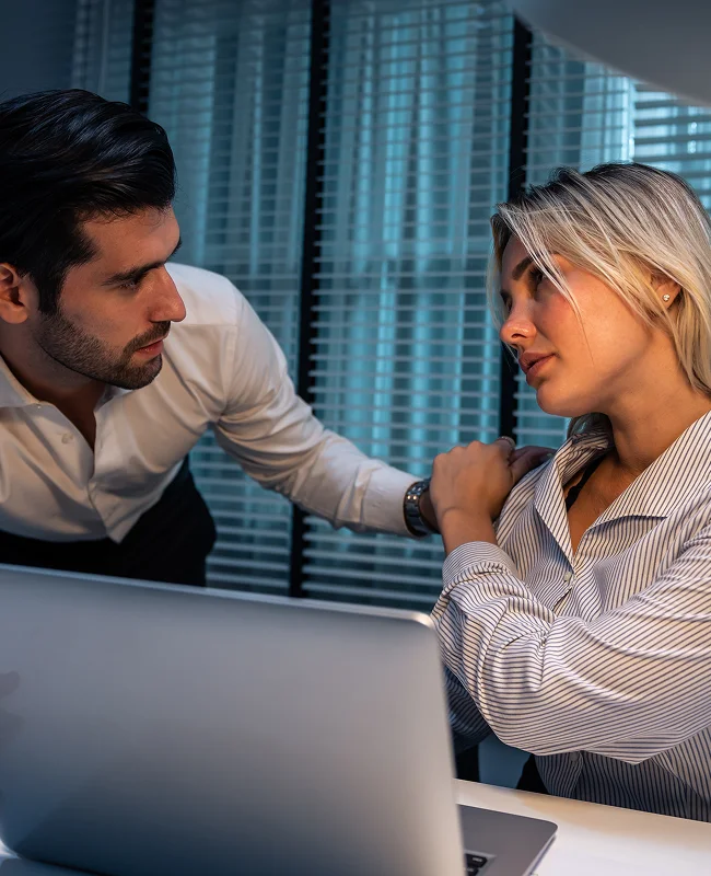 A man in a white shirt leans over a laptop towards a woman in a striped blouse. She looks concerned as he rests a hand on her shoulder in a dimly lit office.