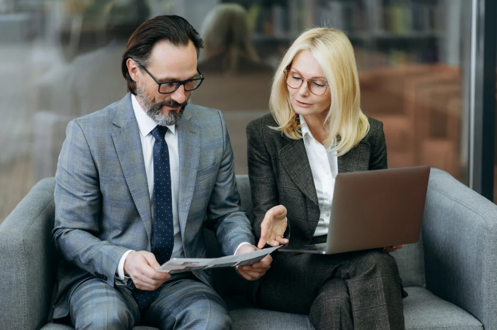 A man in a suit and a woman in formal wear sitting together on a couch, looking at a piece of paper. They both appear to be engaged in discussion, with the man wearing glasses and smiling. A laptop is placed nearby.