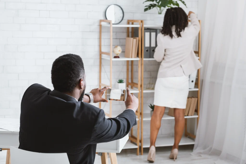 A man in a suit photographs a woman in a pale blazer reaching for a file in a modern office with shelves and plants.
