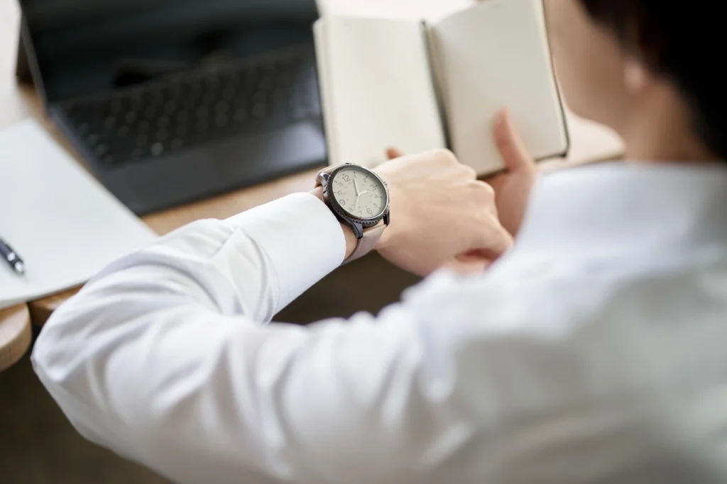 A person in a white shirt checks the time on their wristwatch while holding an open notebook, with a laptop in the background.