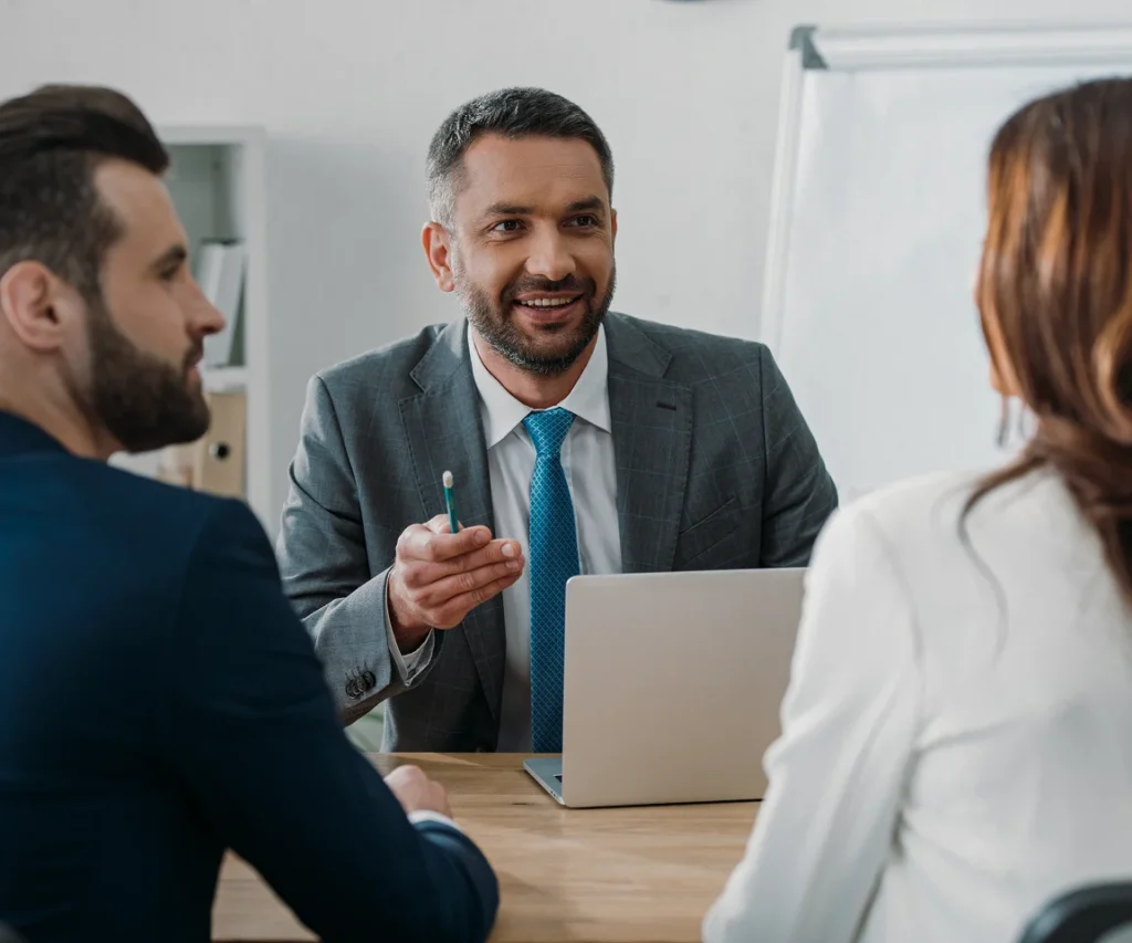 Three people in business attire are having a meeting. A man in a gray suit smiles, gesturing with a pen, while two others listen attentively.