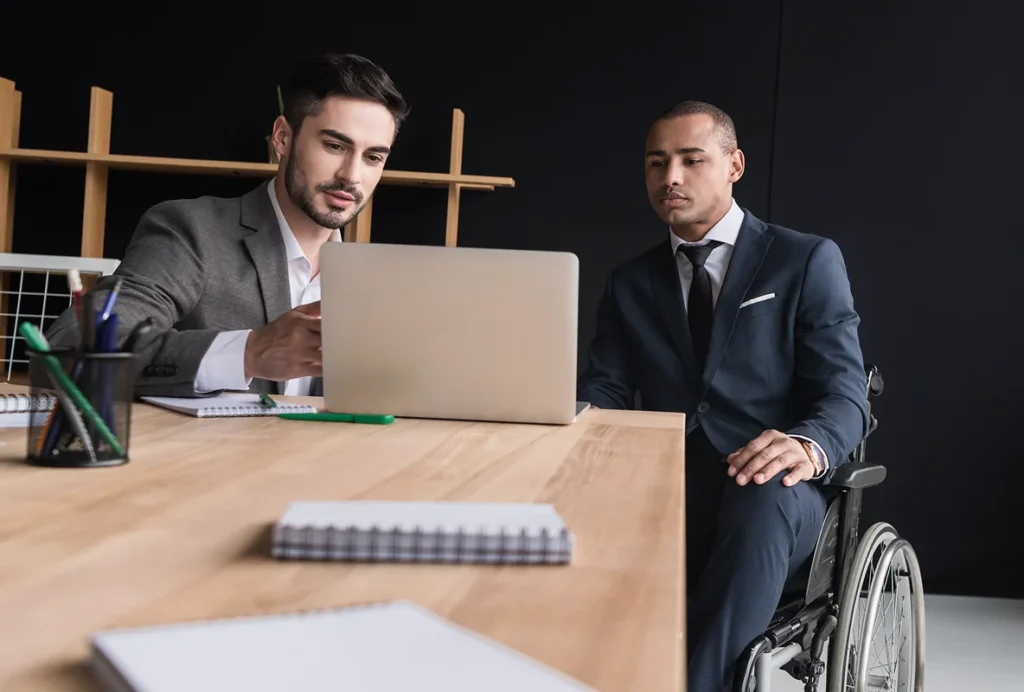 Two men in suits engage in a focused discussion at a desk with a laptop. One man is in a wheelchair, conveying inclusivity in a modern office.